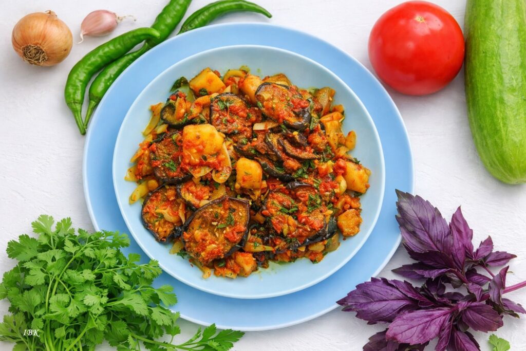 Georgisches Gemüsegericht auf hellblauem Teller mit gebratenen Auberginen, Kartoffeln und Zwiebeln in Tomatensauce, umgeben von frischen Zutaten wie Chili, Knoblauch, Tomate, Gurke, Koriander und violettem Basilikum
